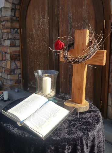 Vor einer verschlossener Kirchentür ist auf einem schwarz eingedeckten Tisch die Bibel, eine Kerze und ein Kreuz mit Dornenkranz und roter Rose aufgebaut.